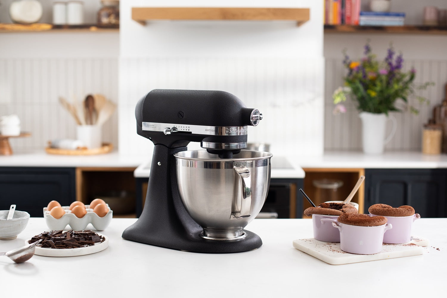KitchenAid Küchenmaschine in Gusseisen Schwarz mit eingesetzter Edelstahlschüssel, daneben stehen kleine Schokoküchlein auf einer weißen Küchenplatte