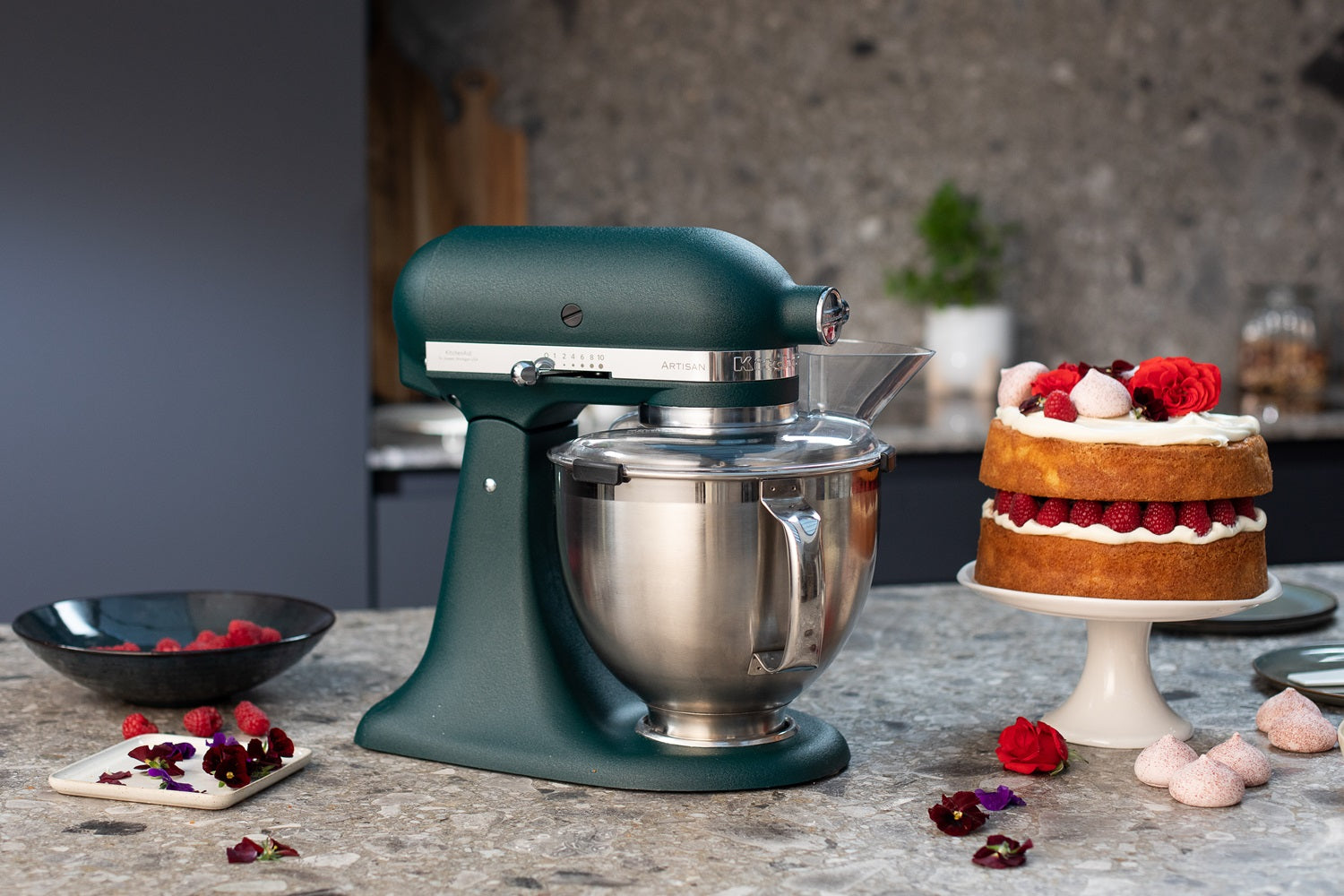 KitchenAid Küchenmaschine in der Farbe pebbled palm mit eingesetzter Schüssel mit Spritzschutz auf grauer Küchenplatte, daneben eine Himbeertorte