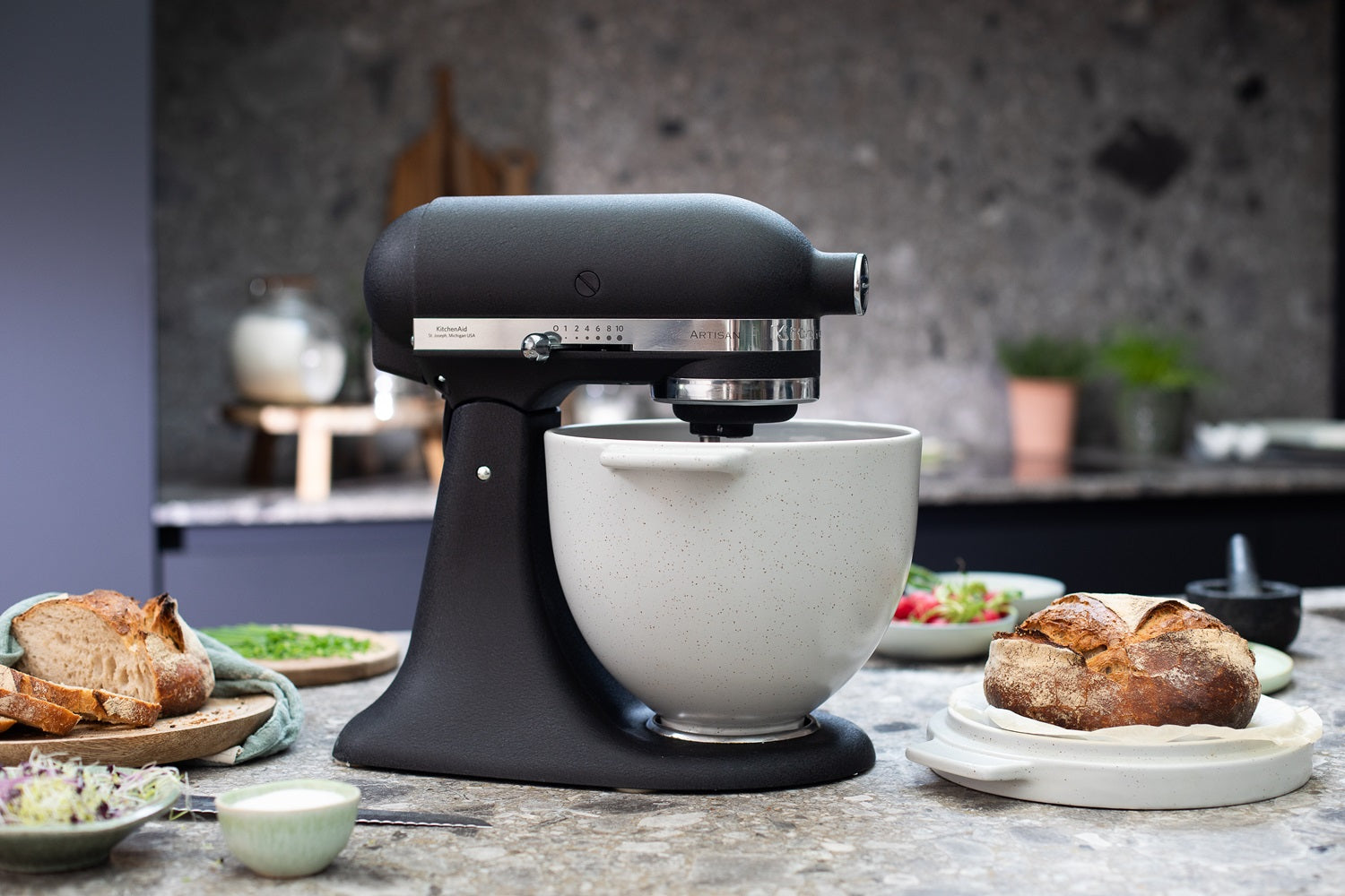 schwarze KitchenAid Küchenmaschine mit eingesetzter Brotbackschüssel, daneben liegt ein Brot auf dem Deckel der Schüssel auf einer hellgrauen Küchenplatte