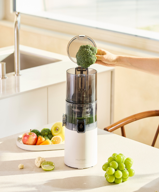 Weißer Hurom-Entsafter in einer hellen Küche, jemand gibt ein Stück Brokkoli in den Einfüllschacht, frisches Obst liegt daneben.