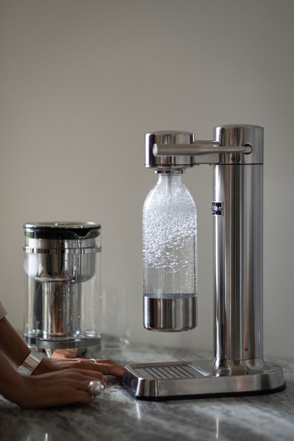 Aarke Wassersprudler in silber sprudelt Wasser auf einer Küchenplatte, daneben wartende Hände