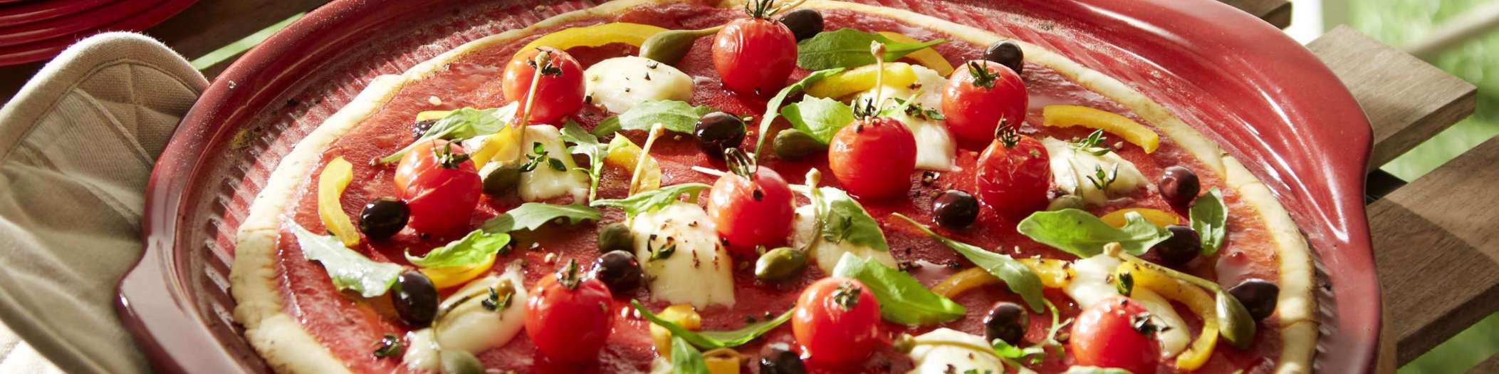 Eine mit Gemüse, Käse und Tomatensauce belegte Pizza liegt auf einem roten Pizzateller von Emile Henry