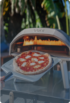 Ooni Pizzaofen im Garten mit Palme im Hintergrund, im Vordergrund eine Pizzaschaufel, auf der eine Pizza liegt