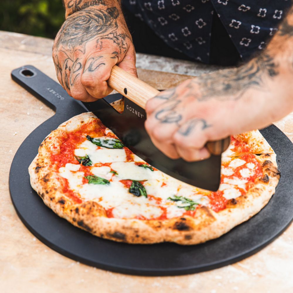 Eine Pizza liegt auf einem Gozney Pizzabrett, man sieht zwei Hände, die mit dem Gozney Pizzarocker die Pizza schneiden