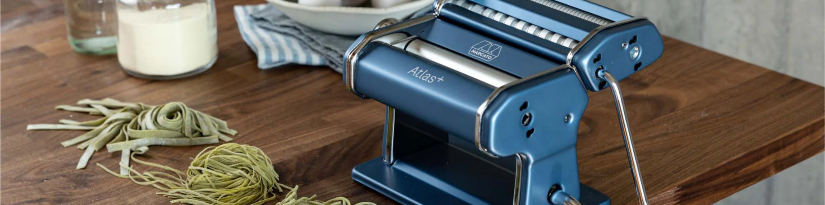 Marcato Atlas + Nudelmaschine in Blau auf einer Küchenarbeitsplatte, daneben frisch hergestellte grüne Pasta, eine Schale mit Eiern und ein Glas mit Mehl