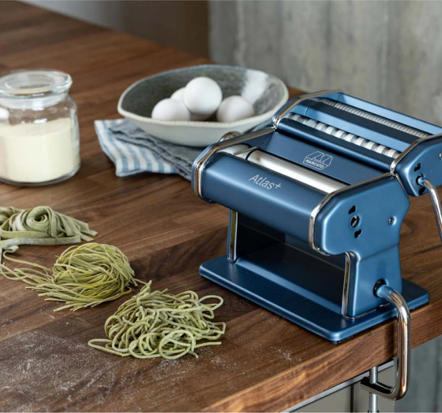 Marcato Atlas + Nudelmaschine in Blau auf einer Küchenarbeitsplatte, daneben frisch hergestellte grüne Pasta, eine Schale mit Eiern und ein Glas mit Mehl