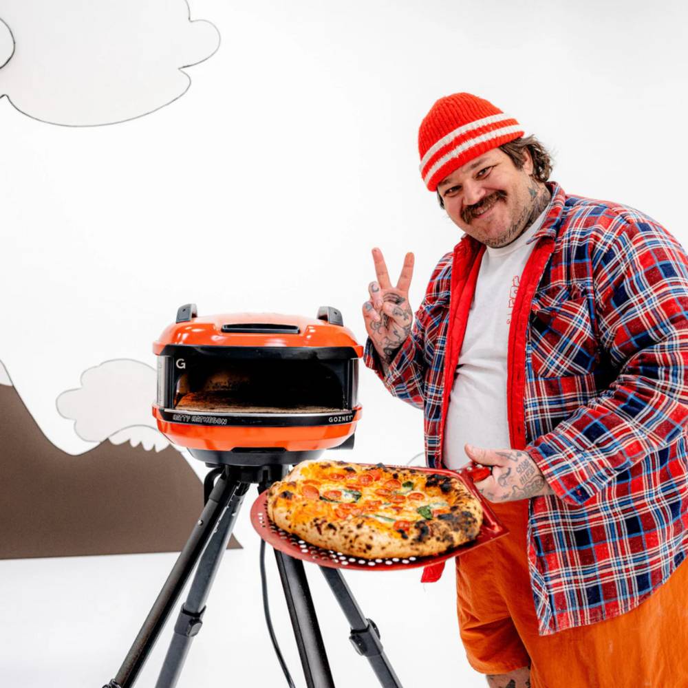 Matty Matheson steht neben dem Gozney Tread in seiner eigenen Special Edition und zeigt das Peace-Zeichen, in der anderen Hand hält er eine Pizza