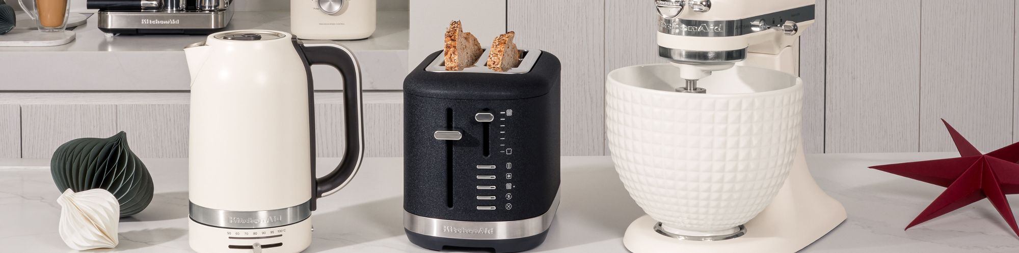 Zusammenstellung verschiedener KitchenAid Produkte. Darunter ein KitchenAid Wasserkocher in der Farbe Weiß, ein KitchenAid Toaster in der Farbe Gusseisen Schwarz sowie eine KitchenAid Artisan Küchenmaschine mit Keramikschüssel in der Farbe Weiß