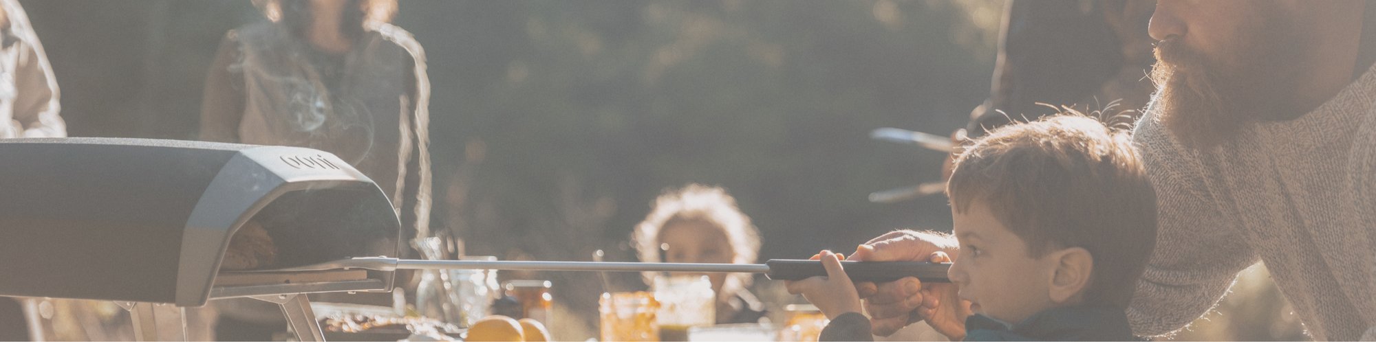 Ein Vater mit seinem Sohn schiebt grade eine Pizza in den Ooni Pizzaofen. Im Hintergrund sind weitere Personen zusehen die vor einem gedeckten Tisch stehen und auf den Pizzaofen schauen.