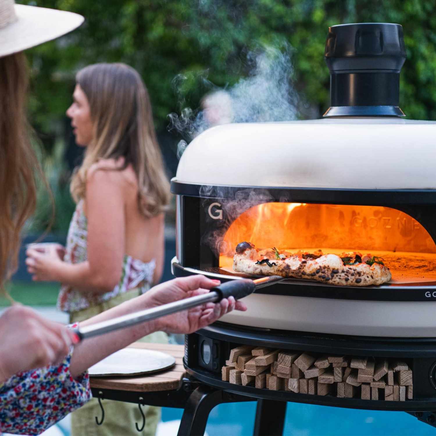 Aufnahme eines Grillabends im Garten mit einer Frau welche einen frisch gebackene Pizza aus einem Pizzaofen der Marke Gozney entnimmt.