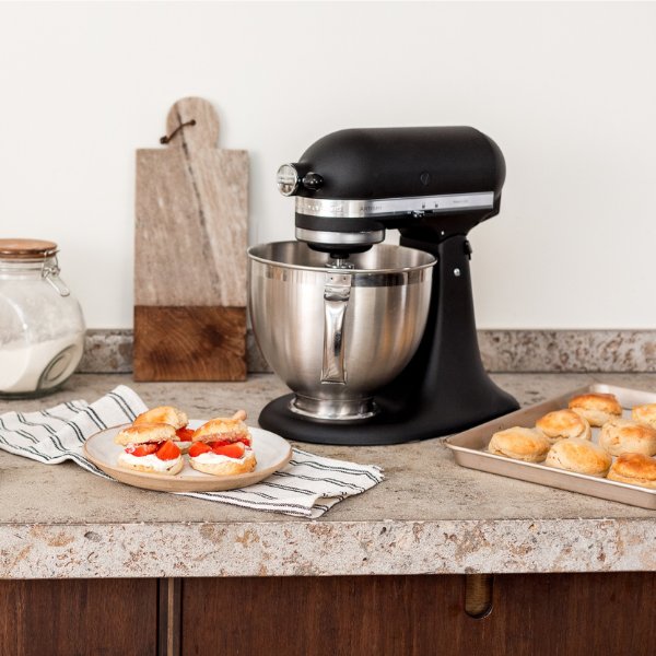 Eine KitchenAid Kuechenmaschine steht auf einer rustikalen Oberfläche in einer modernen Kueche. Vor und neben der Kuechenmaschine stehen fertig zubereitete Muffins.