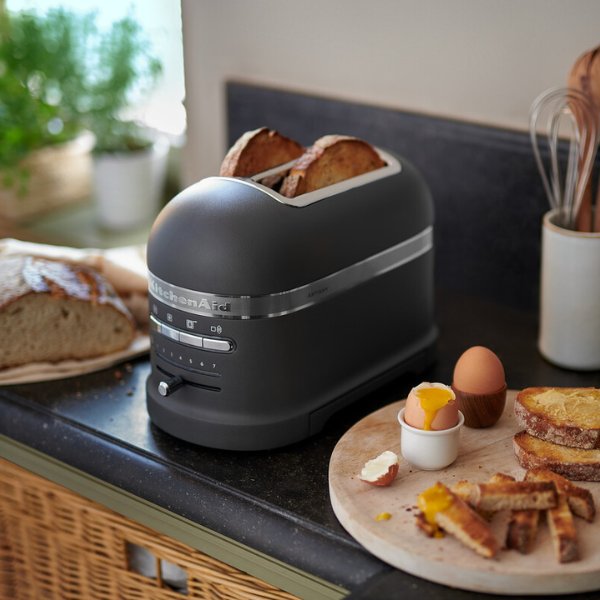 Ein KitchenAid Toaster steht in einer modernen Kueche zusammen mit frisch geschnittenen und getoasteten Brot. In dem Toaster befinden sich zwei fertig getoastete Brotscheiben.