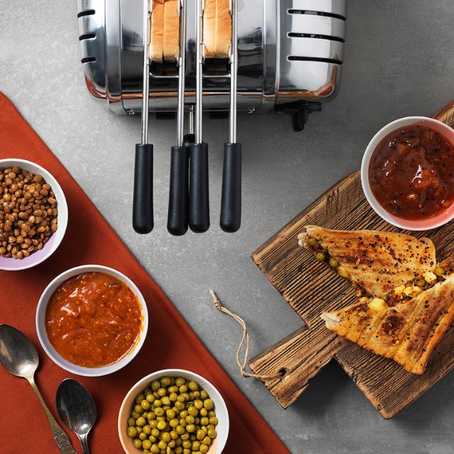 Aufnahme von oben herab auf eine Arbeitsplatte auf der ein Dualit Classic 2er Toaster steht, in welchen 2 Sandwhiches in der Dualit Sandwhichklemme im Toaster stecken. Um den Toaster stehen verschiedene Zutaten und ein Holzbrett mit fertig gegrillten Sandwhiches.