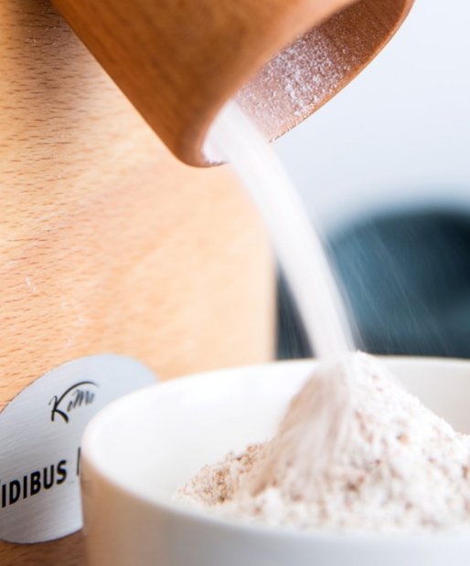 Nahaufnahme einer Komo Fidibus 21 Getreidemühle und einer weißen Keramikschüssel. Aus der Getreidemühle füllt frisch gemalenes Mehl in die weiße Keramikschüssel.