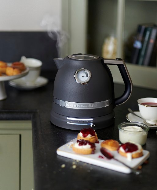 Ein KitchenAid Wasserkocher steht auf einer Kuechenarbeitsplatte, zusammen mit verschiedenen Fruehstuecksgegenständen. Aus dem Wasserkocher steigt verdunstendes Wasser auf.