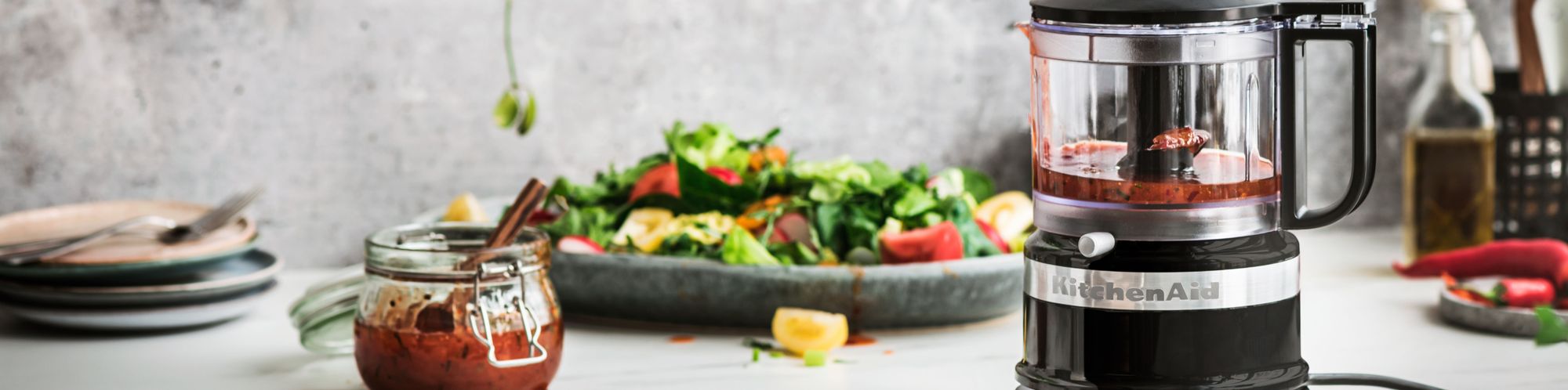 Tischplatte mit einem Salat und selbstgemachten Dressing sowie einer KitchenAid 830 mL Food Processor 5KFC3516 Zerkleinerer in der Farbe Onyx Schwarz, in dem sich Reste des Dressings befinden