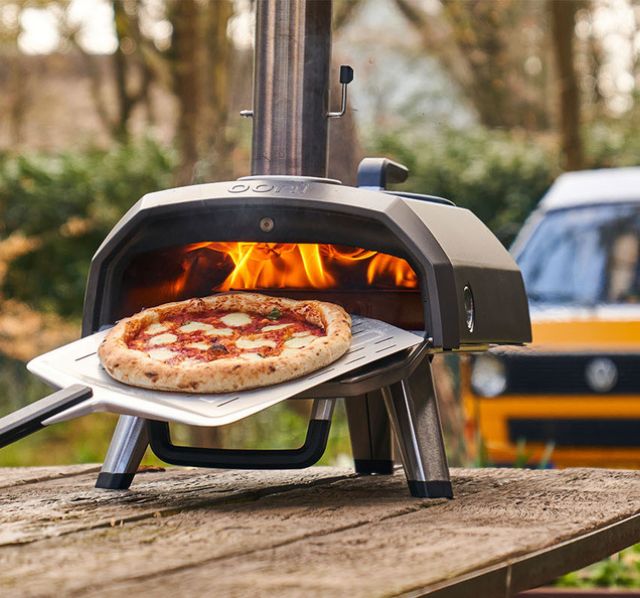 Ein OOni Pizzaofen steht auf einer Picknickbank in der Natur. Aus dem Pizzaofen wird grade eine frisch gebackene Pizza mit einem Pizzaheber herausgeholt.
