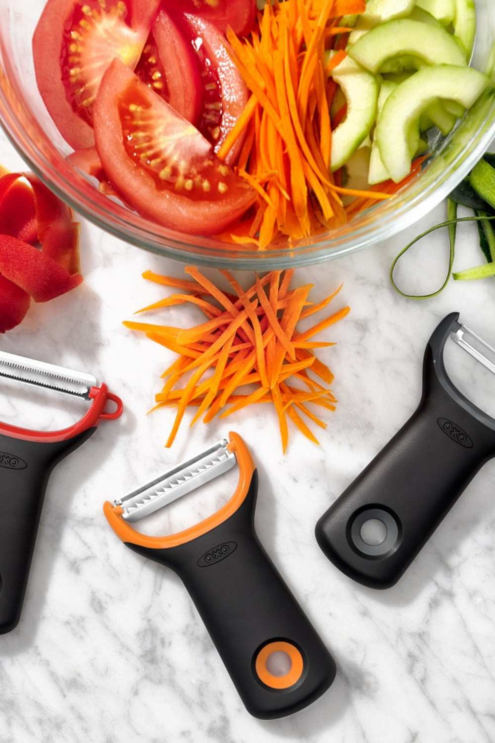 Draufsicht mehrerer OXO Y-Schäler in verschiedenen Farben mit einer Schuessel in dem verschiedenes geschnittenes Gemüse steht