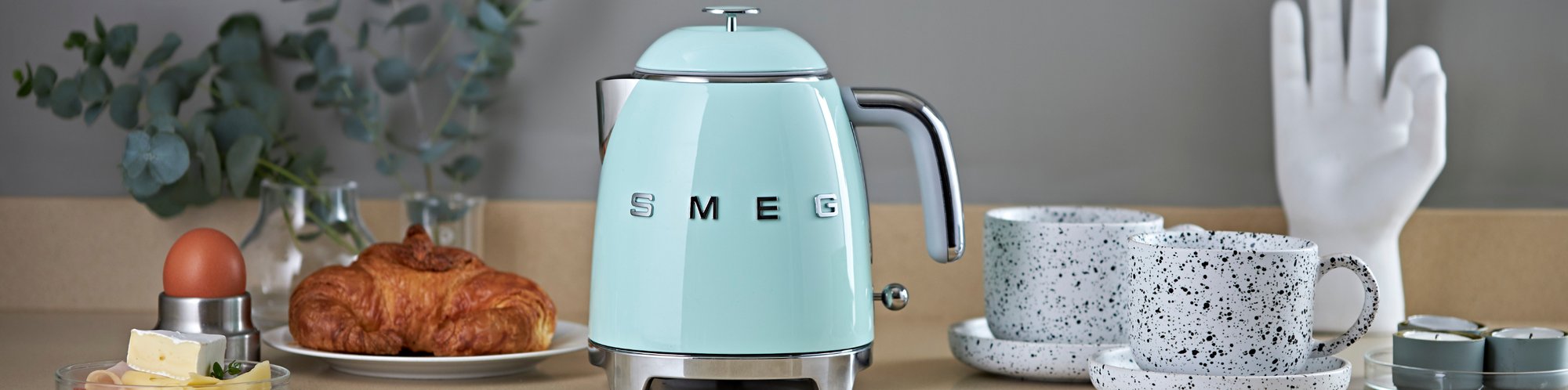 Frontale Ansicht auf einen Smeg 50's Wasserkocher mit 1,7 Liter Fassungsvermögen in der Farbe Pastellblau, wie er zusammen mit zwei Tassen Kaffee, einem Croissant und einem Frühstücksei auf einem Tisch steht
