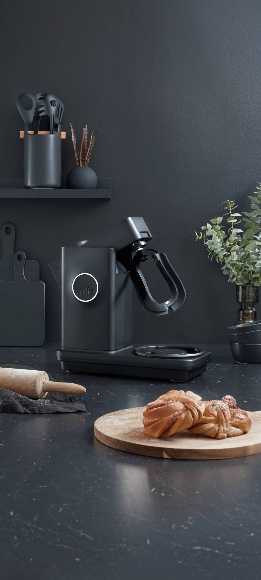 Halbtotale Aufnahme einer WILFA Kuechenmaschine in der Farbe schwarz. De Kuechenmaschine seteht vor einer schwarzen Wand, an der weitere Kuechenutensilien wie Kochlöffel und Schneidebretter hängen