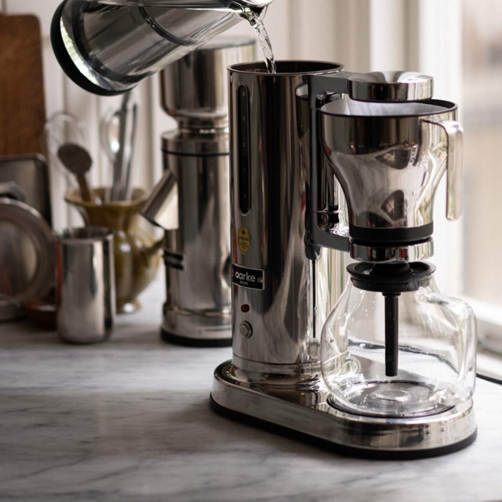 Eine Aarke Kaffeemaschine steht auf einer Marmor Kuechenplatte und wird mit frischem Wasser befüllt. Im Hintergrund sind verschiedene Kuechenutensilien zusehen.