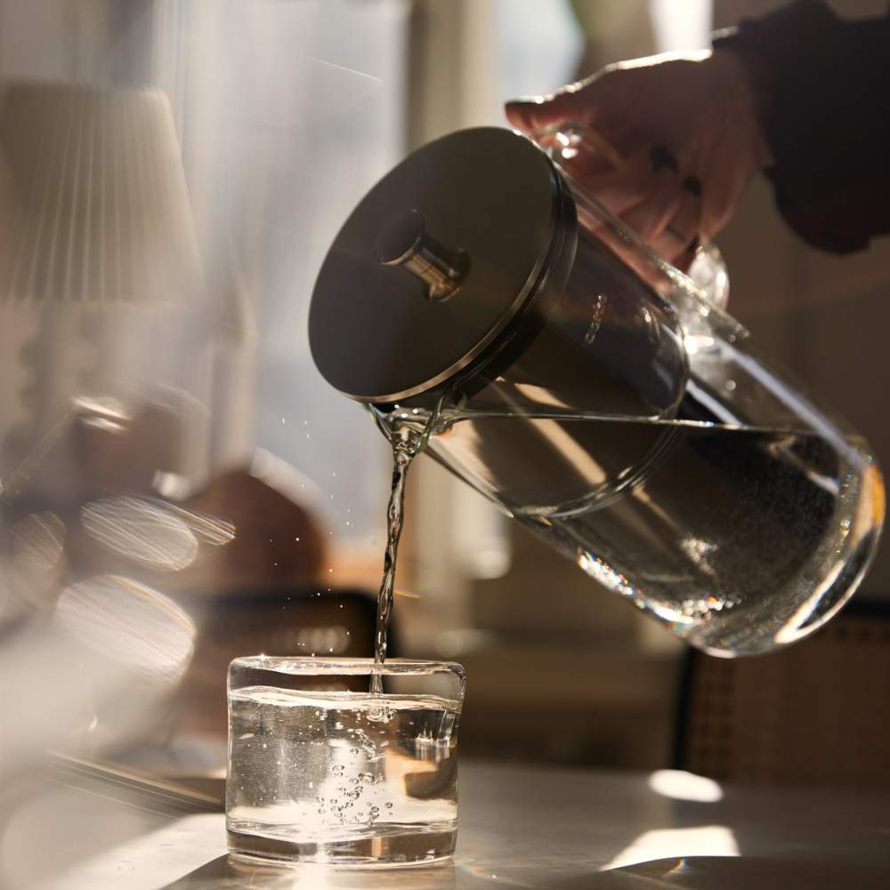 Mit einem Wasserfilter der Marke Aarke wird gefiltertes Wasser in ein Glas, welches auf einem Tisch steht, gegossen.