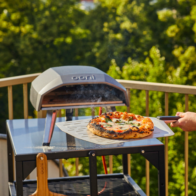 Frisch gebackene Pizza auf Pizzaschieber vor Ooni Pizzaofen stehend auf Ooni Modultisch auf der Terrasse
