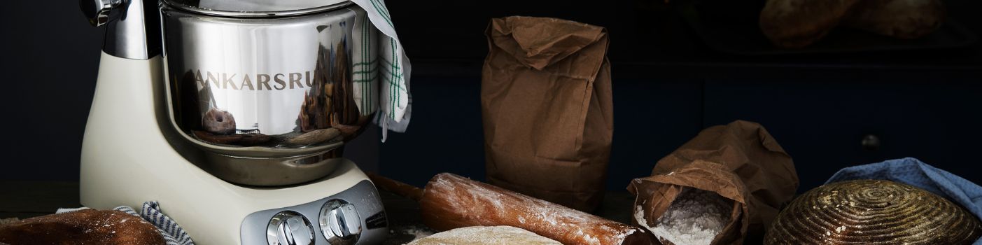 Küchenmaschine von Ankarsrum auf bemehlter Arbeitsfläche mit Broten, Mehl und Backutensilien.