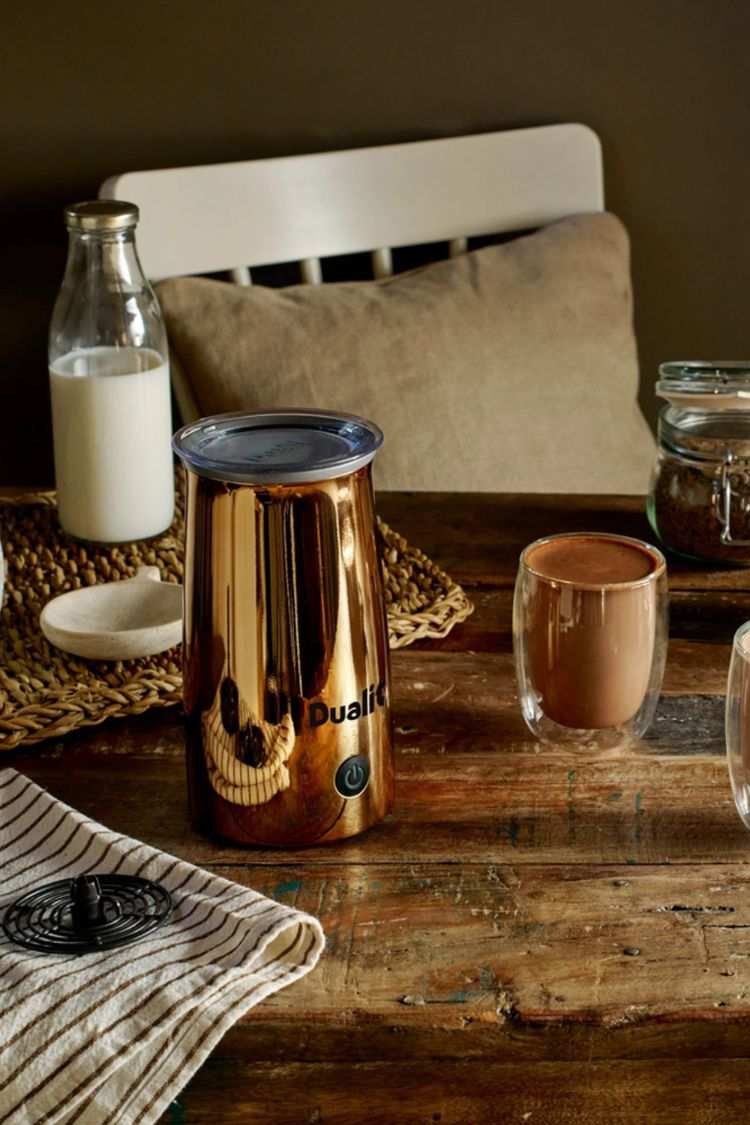 Ein Dualit Milchaufschäumer steht auf einem rustikalen Holztisch zusammen mit einem Glas Kakao, frischer Milch und einer Tasse.