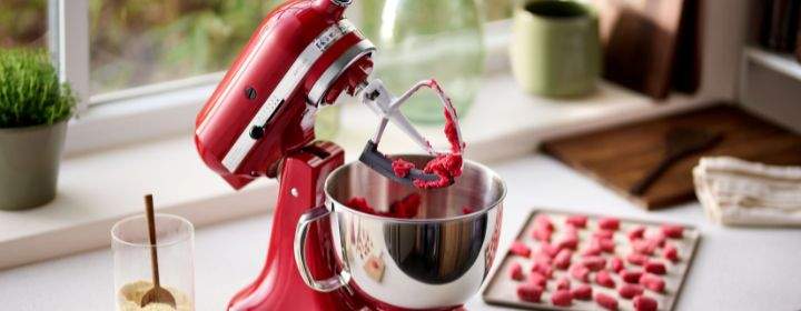 KitchenAid Kuechenmaschine steht auf einer Kuechenarbeitsplatte. In der Schuessel befindet sich roter Teig, wovon man im Hintergrund auch schon fertige Zubereitungen sieht.