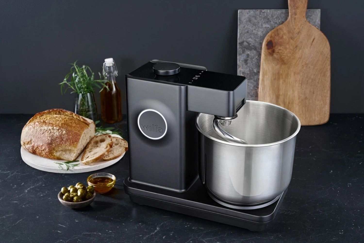 Eine Wilfa Kuechenmaschine steht auf einer Arbeitsplatte mit frisch geschnittenen Brot, Oliven, Olivenöl und Schneidebretter.