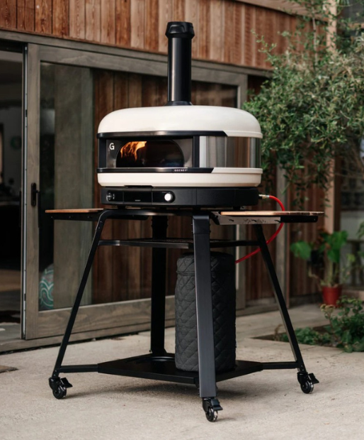 Moderner Outdoor-Pizzaofen auf stabilem Metallgestell mit Holzablagen, betrieben mit Gas, vor einem Haus mit Terrassentüren.