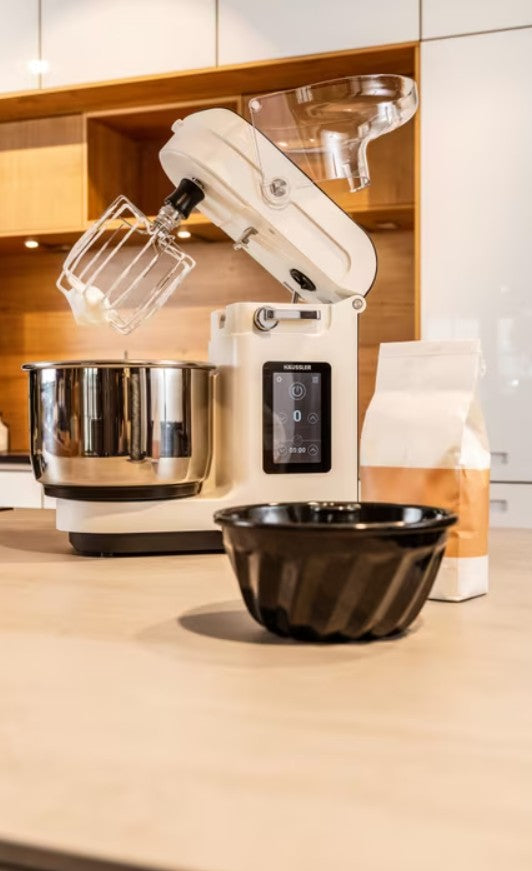 Weiße Häussler Küchenmaschine mit Rührbesen und Edelstahlschüssel auf einer Küchenarbeitsfläche, daneben eine Schüssel und eine Packung Mehl.