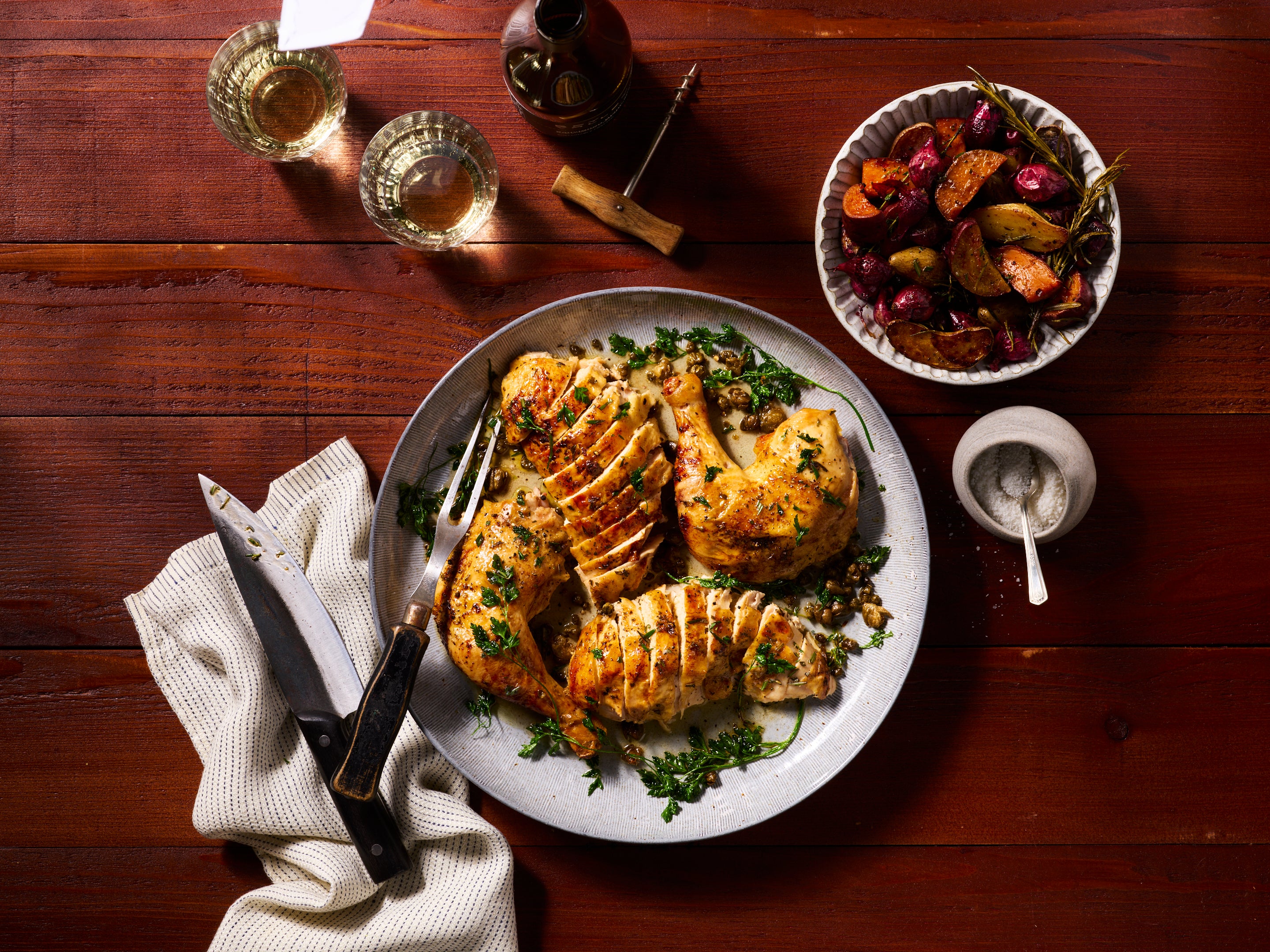 Von oben fotografierter Teller mit aufgeschnittenem, gebratenem Hähnchen und frischen Kräutern auf einem Holztisch, daneben Besteck, ein Tuch, zwei Gläser Weißwein sowie eine Schale mit geröstetem Gemüse.