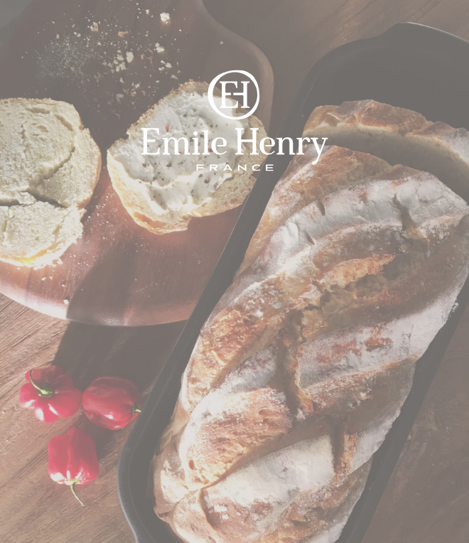 Frisch gebackenes Brot in einer Emile Henry Backform, daneben Brotscheiben mit Aufstrich auf einem Holzbrett, dekoriert mit roten Chilis, präsentiert mit Emile Henry Logo
