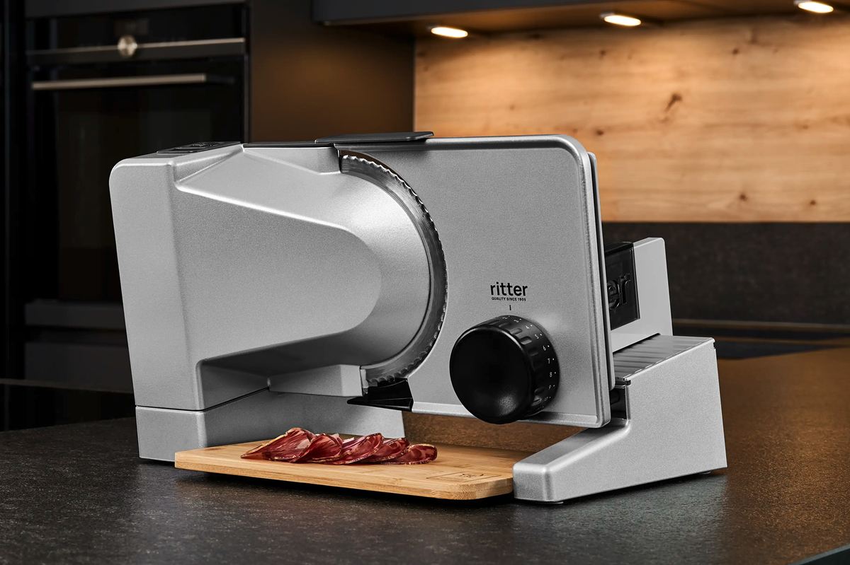 Elektrische Allesschneidemaschine von Ritter aus Metall auf einer Küchenarbeitsfläche, mit dünn geschnittenen Scheiben Wurst auf einem Holzbrett davor.
