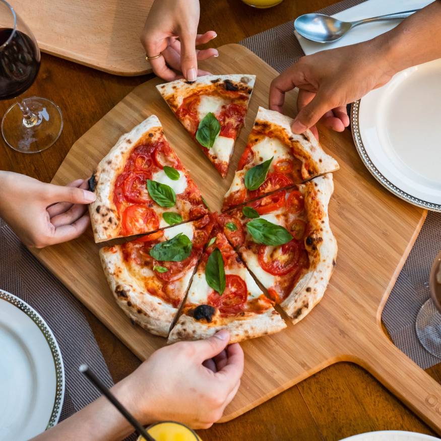 EIne Pizza liegt in 6 Teile geschnitten auf einem Ooni Pizzabrett aus Holz, vier Hände nehmen sich je ein Stück