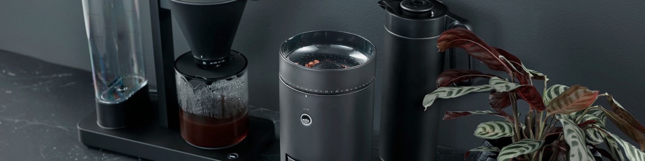 Wilfa Kaffeemühle, Kaffeemaschine und Thermoskanne in schwarz auf schwarzer Oberfläche und vor schwarzer Wand, eine grüne Blume als Farbtupfer