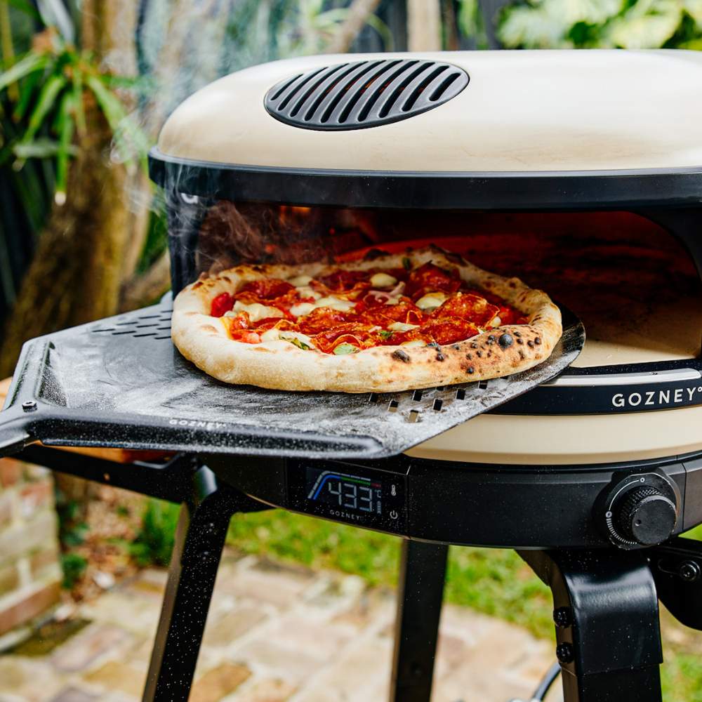 Nahaufnahme des Arc Pizzaofen in Cremefarben von der Marke Gozney, wie eine Pizza auf einem Pizzaschieber aus der Öffnung gezogen wird.