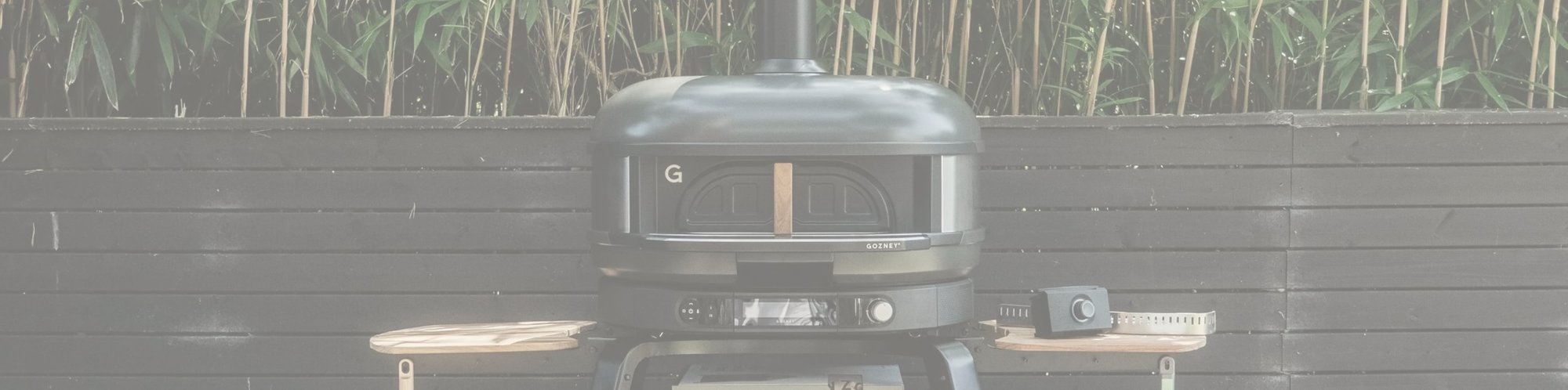 Leicht ausgegrautes Panorama Bild des Gozney Dome XL Pizzaofen in der Farbe "Off Black" vor einem Gartenzaun stehend.