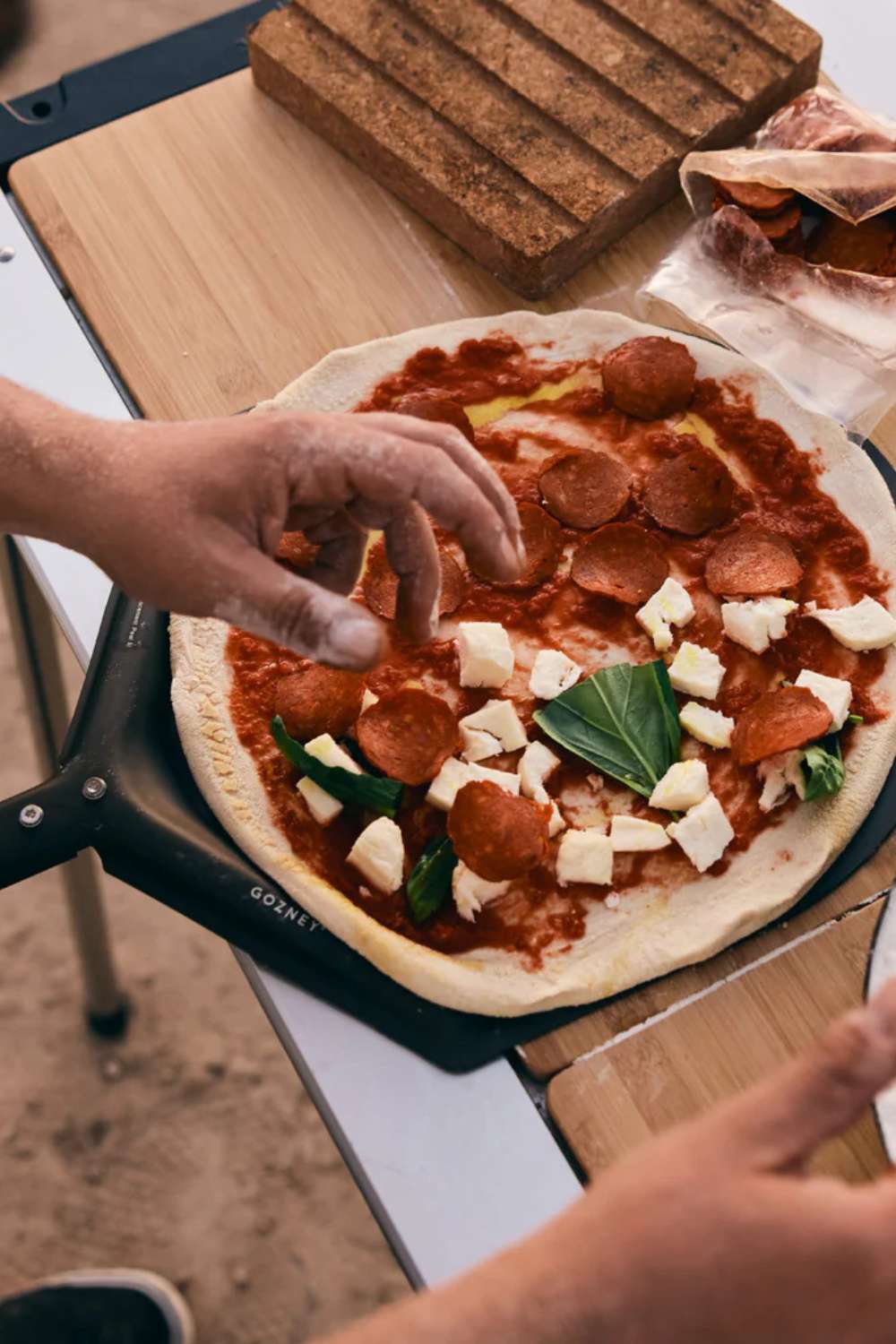Aufsicht auf einen frisch ausgerollten Pizzateig, welcher auf einem Gozney Pizzaschieber liegt und von einer Person mit Toppings beleget wird.