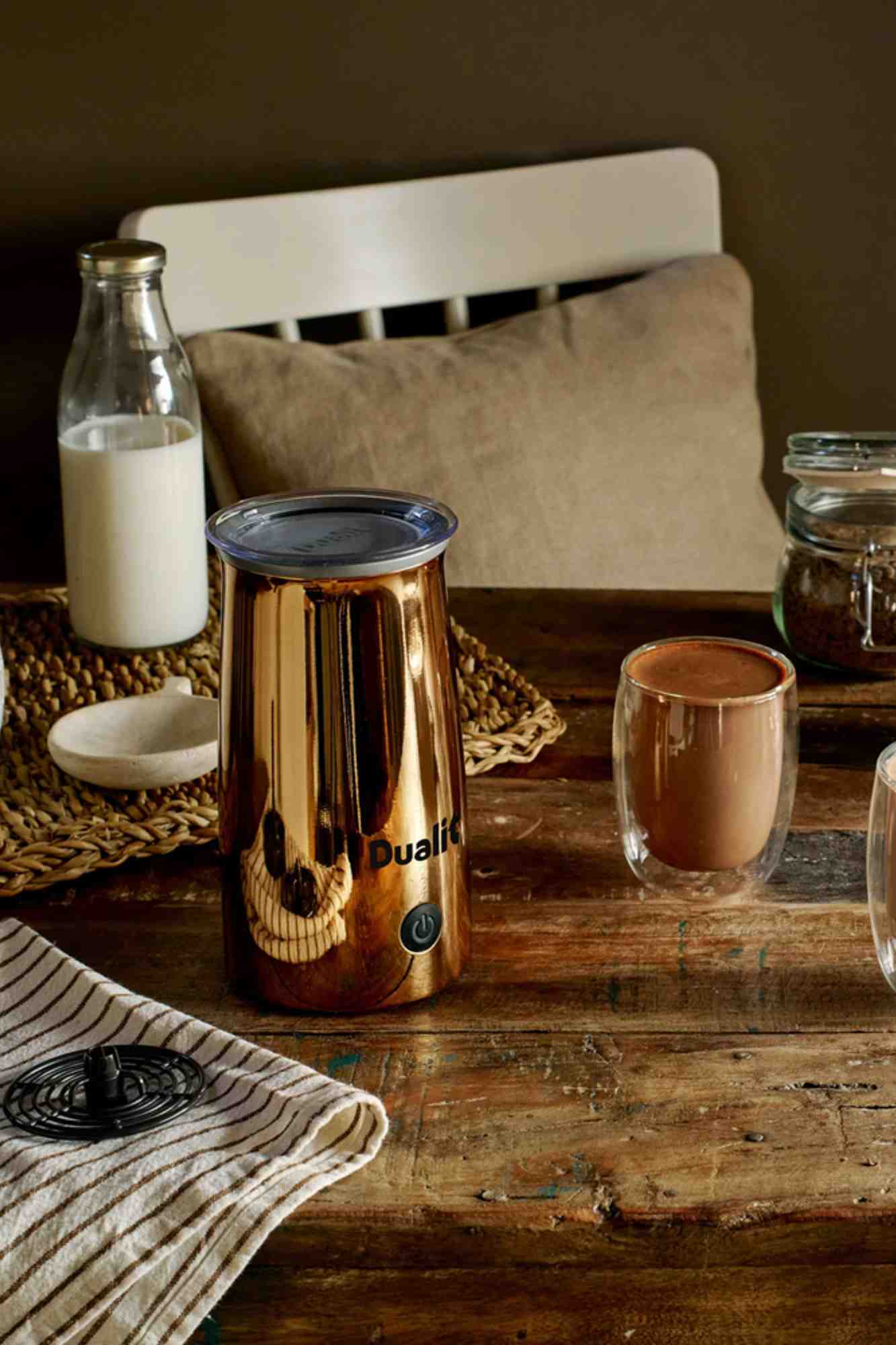 Ein Dualit Milch- und Schokoladenaufschäumer in der Farbe Kupfer steht auf einem rustikalen Holztisch neben einem Glas Kakao und einer Flasche Milch.