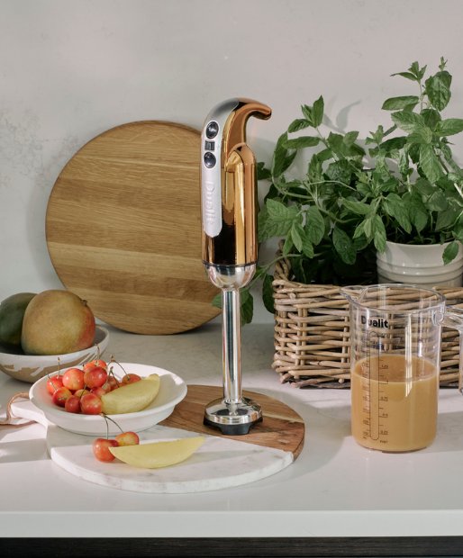Aufnahme einer Kueche in dessen Mitte ein Dualit Stabmixer in der Farbe Kupfer auf einem Holzschneidebrett steht. Neben dem Stabmixer befindet sich eine Schuessel mit Kirschen und weiteren Früchten sowie einem Dualit Messbecher, welcher mit Frisch zubereiteten Fruechtepueree gefüllt ist.