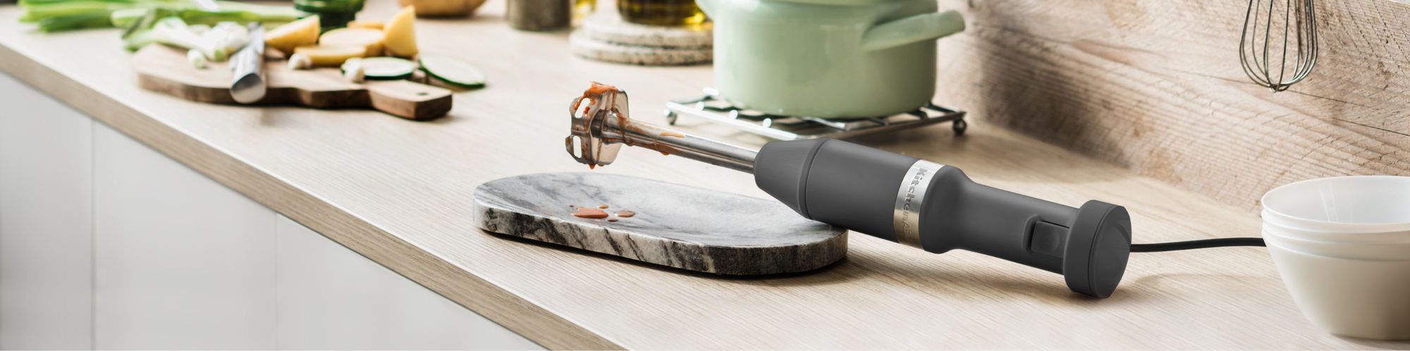 Benutzter KitchenAid Kabel-Stabmixer 5KHBV83E in der Farbe Schwarz matt, liegend auf einem Holzkuechentisch, zusammen mit anderen Zutaten