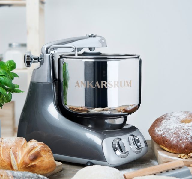 Ein gedeckter Tisch auf dem Mittig eine Ankarsrum Kuechenmaschine steht. Um die Kuechenmaschine herum befinden sich zwei Schneidebretter auf denen frisch gebackenes Brot befindet.