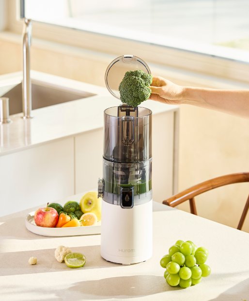 Ein Hurom Slow Juicer E30ST steht auf einem weißen Kuechentisch, in einer Kueche. Auf dem Tisch liegen verschiedene Obst und Gemüsesorten.