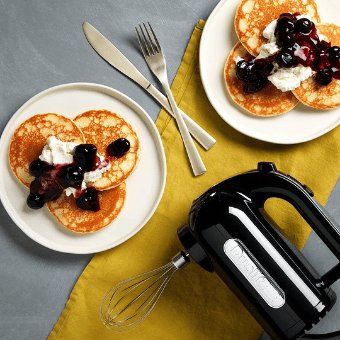 Ein gedeckter Tisch mit zwei Tellern, Besteck und einem Dualit Handmixer, welcher auf einem gelben Kuechentuch liegt. Auf den Teller liegen frisch zubereitete Pfannkuchen mit verschiedenen Toppings.