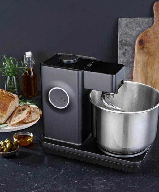 Eine Wilfa Kuechenmaschine steht auf einer schwarzen Kuechenarbeitsplatte vor grauen Hintergrund. Neben der Kuechenmaschine liegt ein frisch gebackenes Brot sowie eine Schuessel mt Olivenöl und eingelegten Oliven.