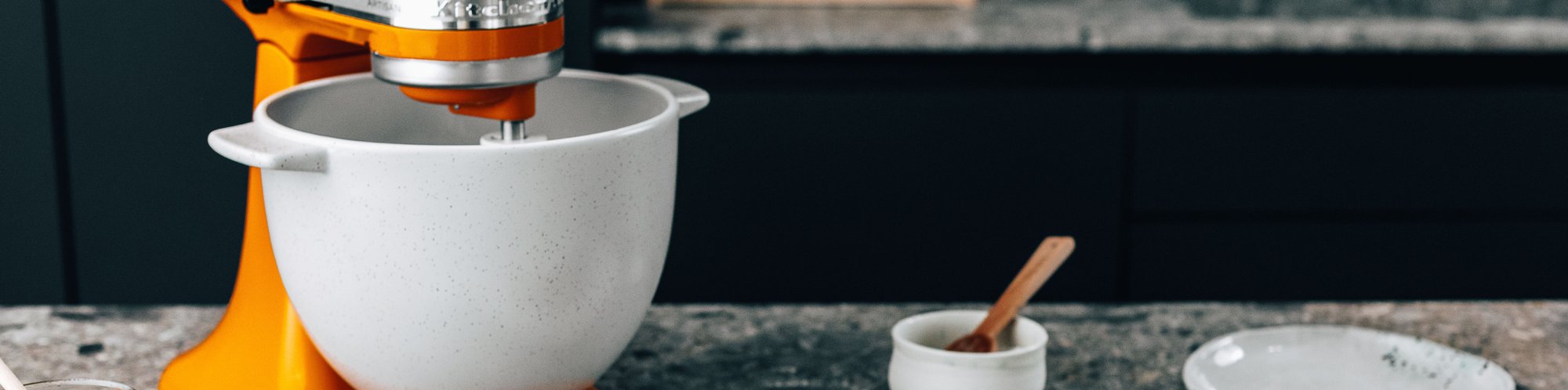 Nahaufnahme einer KitchenAid Kuechenmaschine in der Farbe Orange, stehend auf einem Tisch mit einem Teller sowie einem Gewürzglas