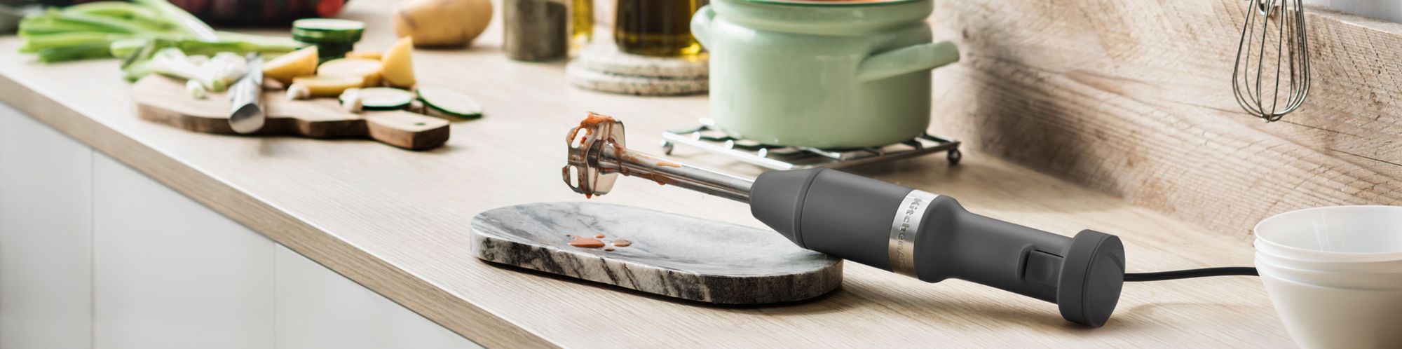 Benutzter KitchenAid Kabel-Stabmixer 5KHBV83E in der Farbe Schwarz matt, liegend auf einem Holzkuechentisch, zusammen mit anderen Zutaten